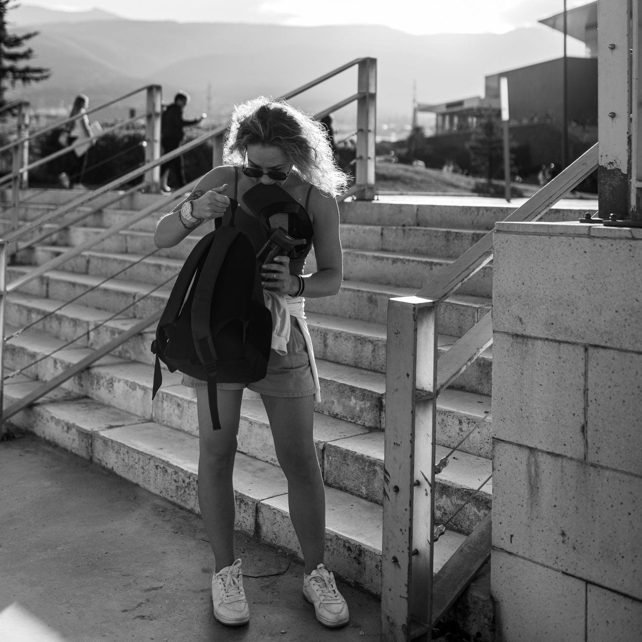 Автентична черно-бяла снимка, показваща способността на фотографа да улавя истински моменти. Силната задна светлина (контражур) е използвана креативно, за да подчертае фигурата и да добави атмосфера към този непринуден градски кадър, докато жената търси в раницата си.
