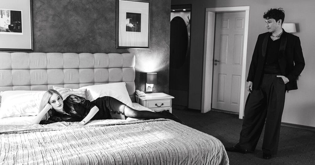 A woman lying on a bed and a man standing in a hotel room.