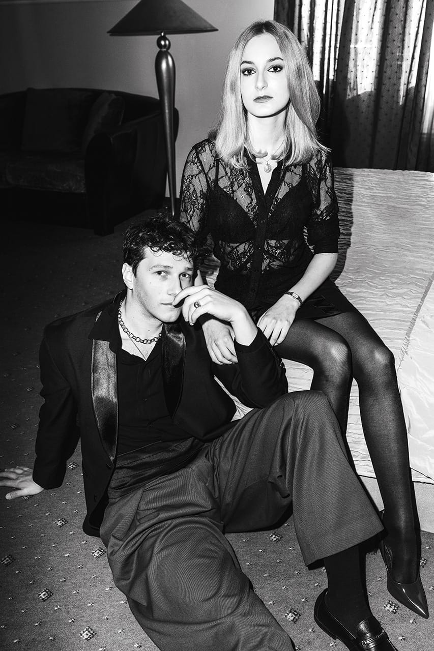 A man sitting on the floor and a woman sitting on a bed behind him, both looking at the camera, in black and white.