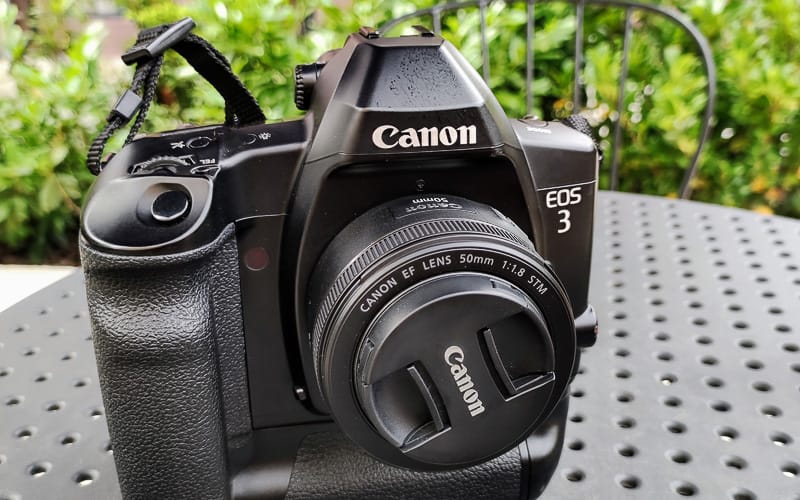 Black Canon EOS 3 film camera with a 50mm f/1.8 STM lens and lens cap on a dark metal table outdoors.