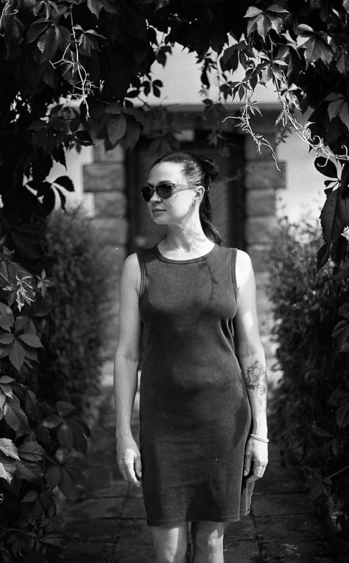 Black and white portrait of a woman in sunglasses and a sleeveless dress, standing under leafy vines on a garden path.