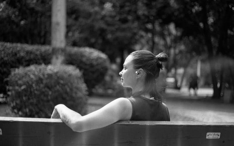 Black and white film photo of a woman with her hair in a bun, sitting on a park bench and looking to the left, captured on Kentmere 400 film.
