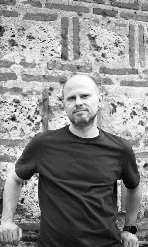 Black and white portrait of a bearded man with a direct gaze, leaning against an old, textured brick wall.