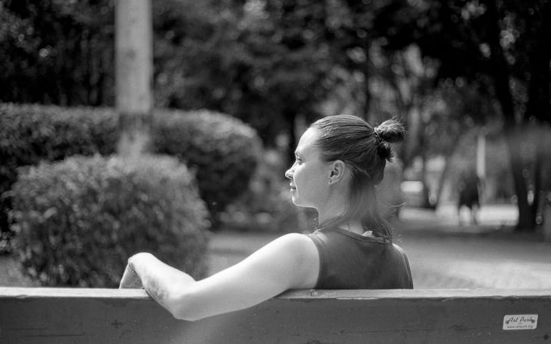 A woman with her hair in a bun sits on a park bench, seen from behind and to her left, looking into the blurred park background.